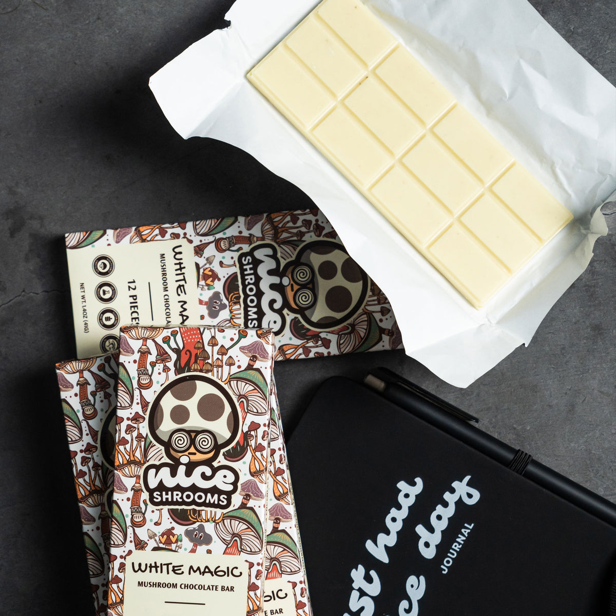 A styled flat-lay of a white chocolate bar and its packaging on a dark gray surface. The main product is a white chocolate bar partially unwrapped, alongside a printed box labeled 'White Magic Mushroom Chocolate Bar' by 'Nice Shrooms' and a black journal with white text.