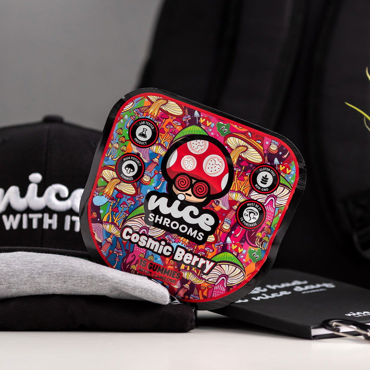 A square product package labeled 'nice SHROOMS Cosmic Berry Gummies' with a highly colorful psychedelic mushroom illustration, styled on a tabletop with black branded merchandise in the background.