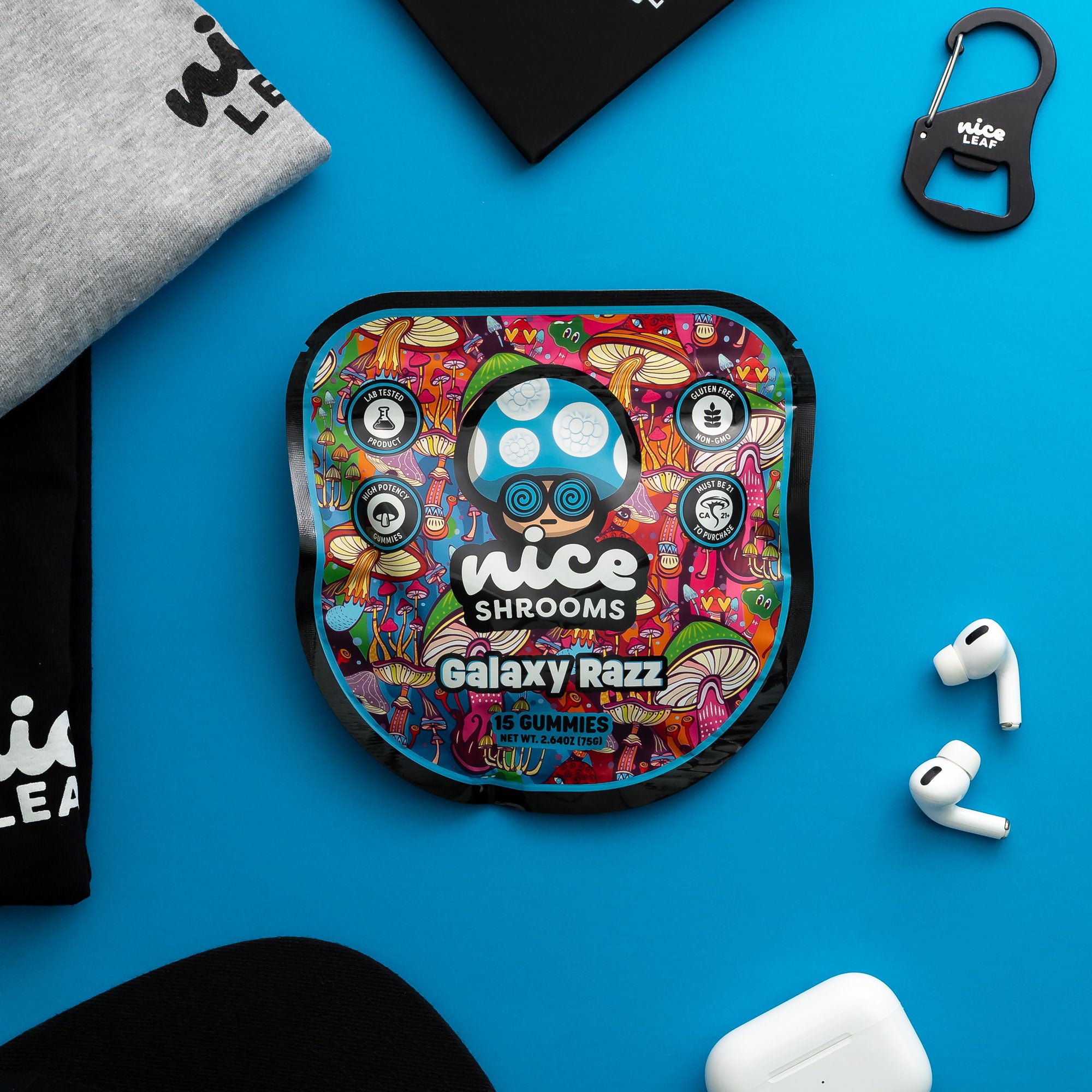 A colorful round case for Galaxy Razz gummies by Nice Shrooms, styled in a flat-lay on a bright blue background with a gray sweatshirt, black t-shirt, carabiner, wireless earbuds, and a charging case.