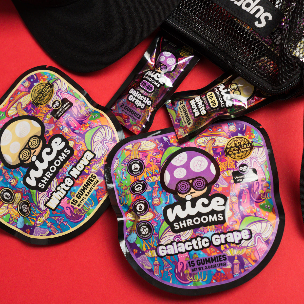 Two nice Shrooms Galactic Grape gummy pouches and two individual gummy packets are arranged on a red surface next to a black cap and a black mesh pouch. The packaging features a highly colorful, psychedelic space-themed design with a purple shroom character.