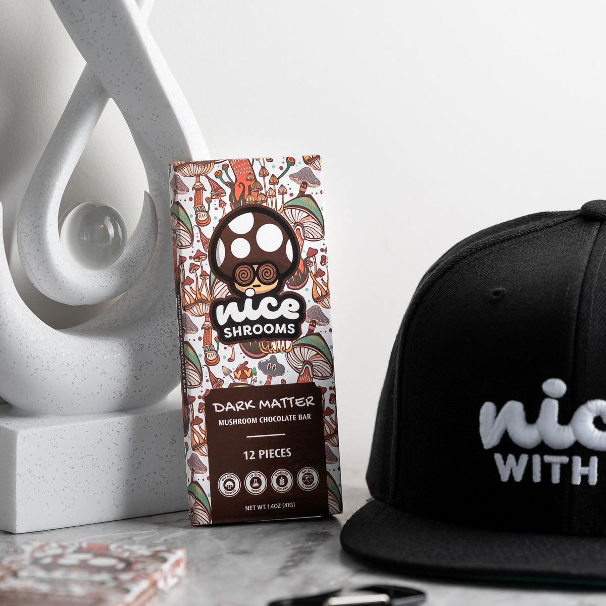 A package of Nice Shrooms Dark Matter Mushroom Chocolate Bar stands on a tabletop next to a black baseball cap with white 'nice WITH' lettering and a white decorative sculpture in the background.