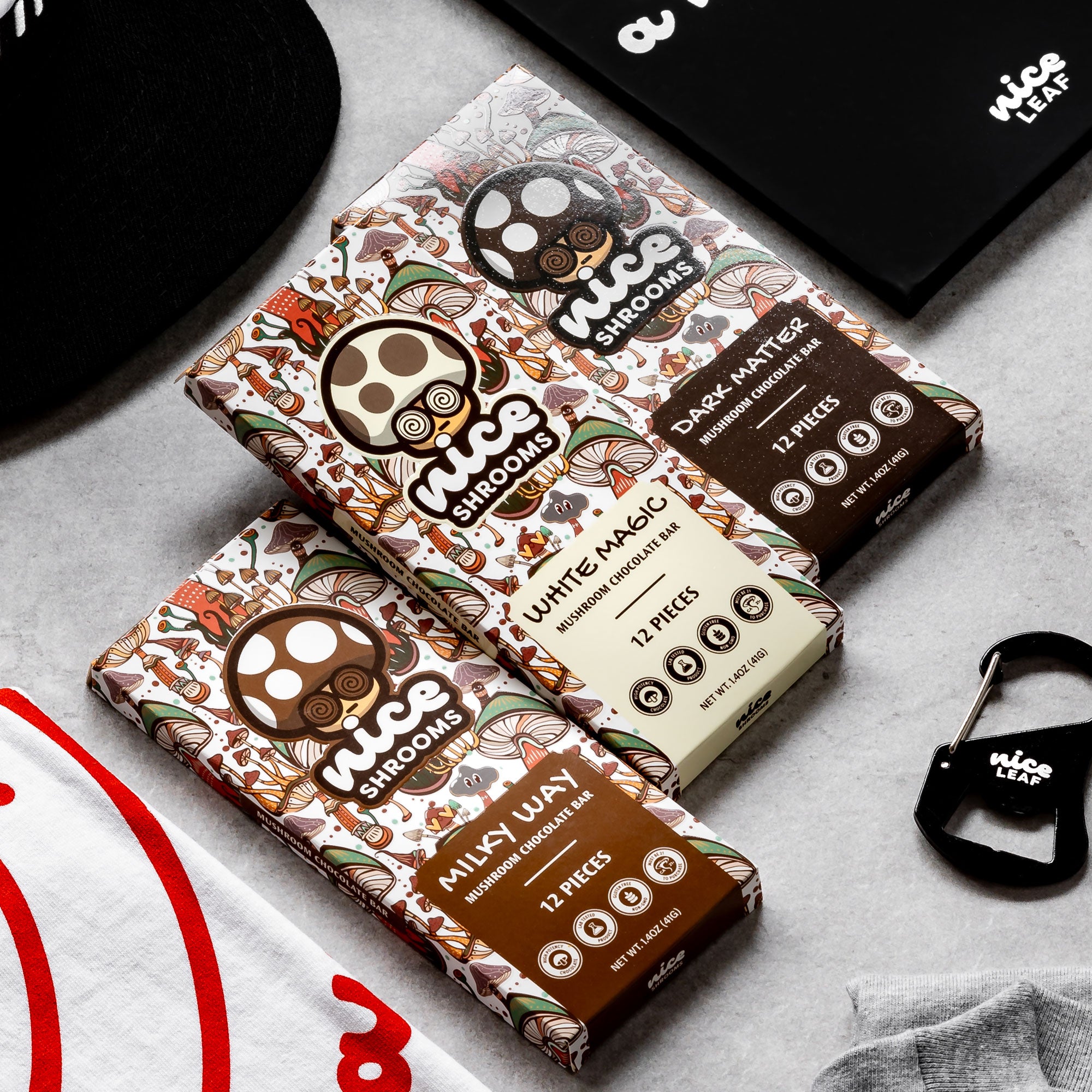 Three packaged chocolate bars from Nice Shrooms arranged on a light gray surface with a black cap, black notebook, and small black keychain nearby. The packages feature colorful owl illustrations and labels for Dark Chocolate, White Chocolate, and Milk Chocolate.