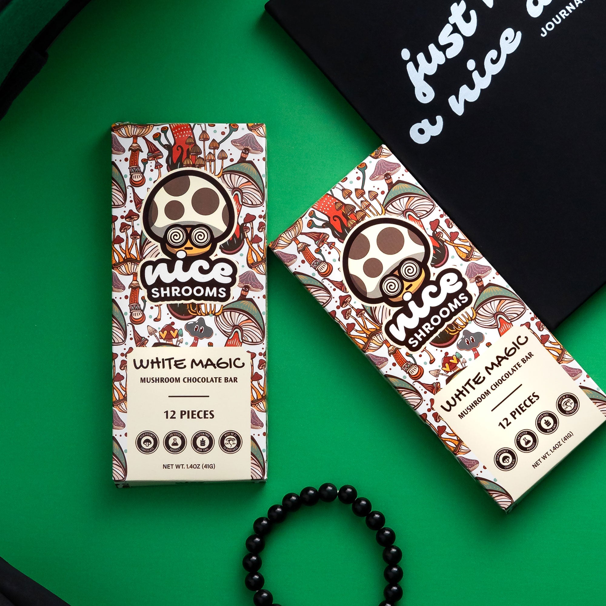 Two boxes of Nice Shrooms White Magic Mushroom Chocolate Bar arranged on a bright green background with a black journal and a black beaded bracelet as props.