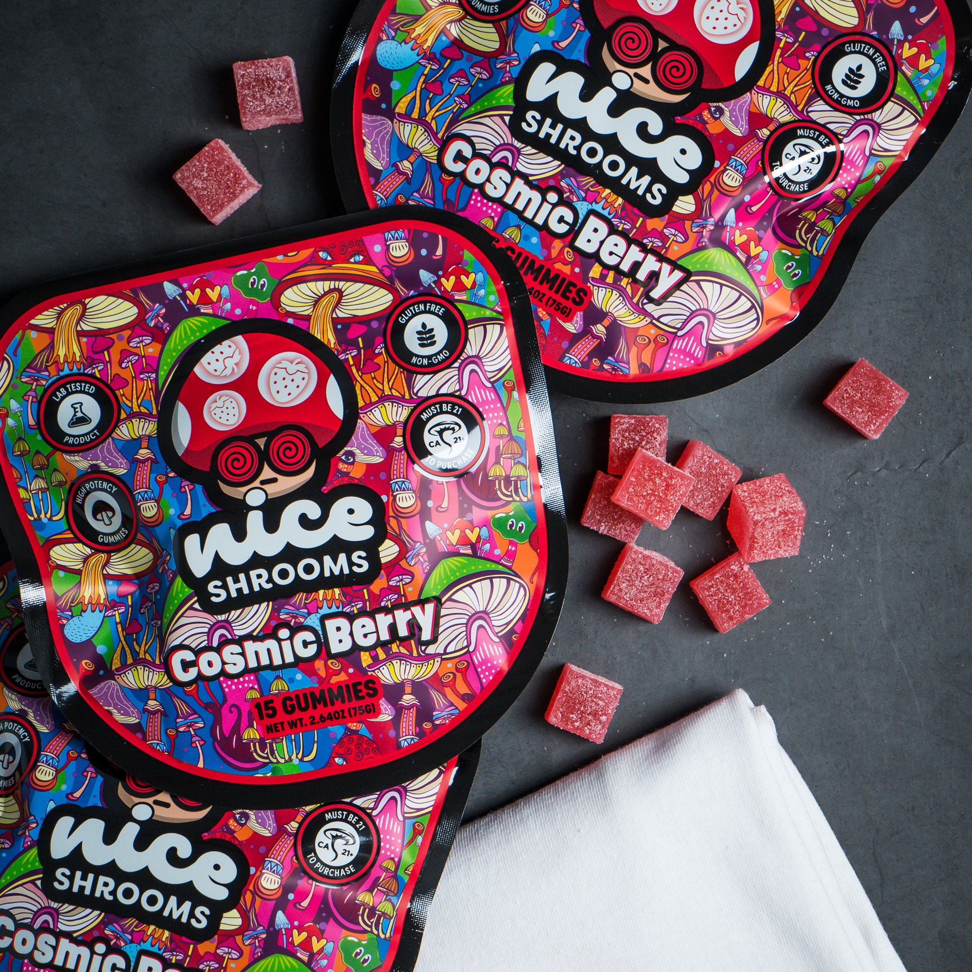 Several colorful round packages of Nice Shrooms Cosmic Berry gummies are arranged on a dark surface, with scattered pink-red gummy cubes and a folded white cloth visible. The packaging has a highly detailed psychedelic multicolor design with the brand name 'nice SHROOMS' and product name 'Cosmic Berry' clearly visible.