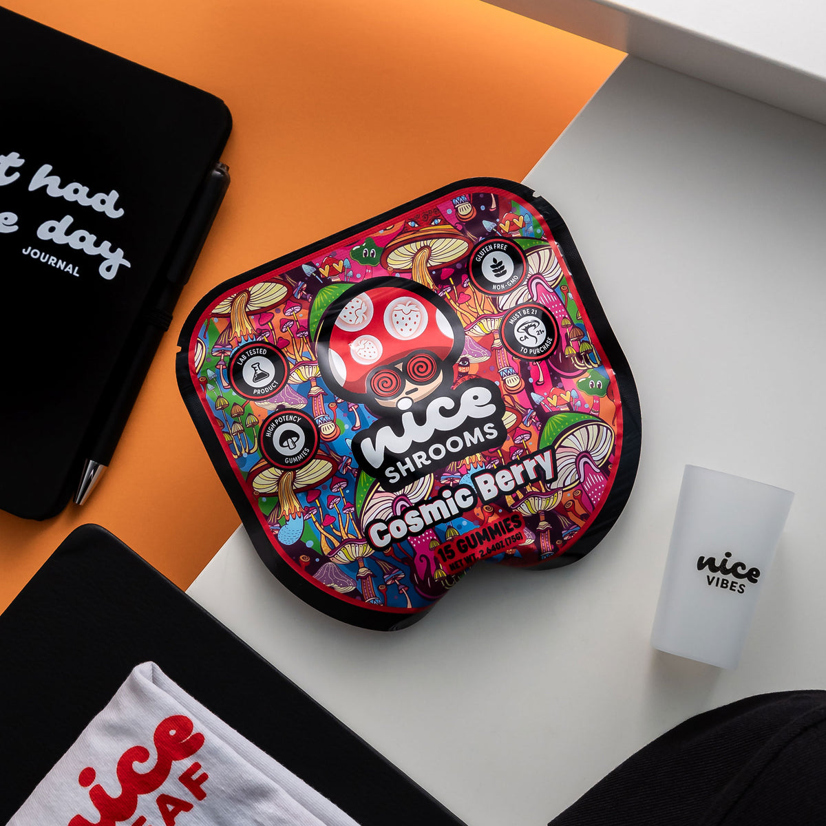 A colorful, mushroom-shaped pouch with a psychedelic cartoon design and the text 'nice SHROOMS Cosmic Berry' is displayed on a white surface next to a white cup, a black notebook, and a white t-shirt. The background is split diagonally between orange and white.