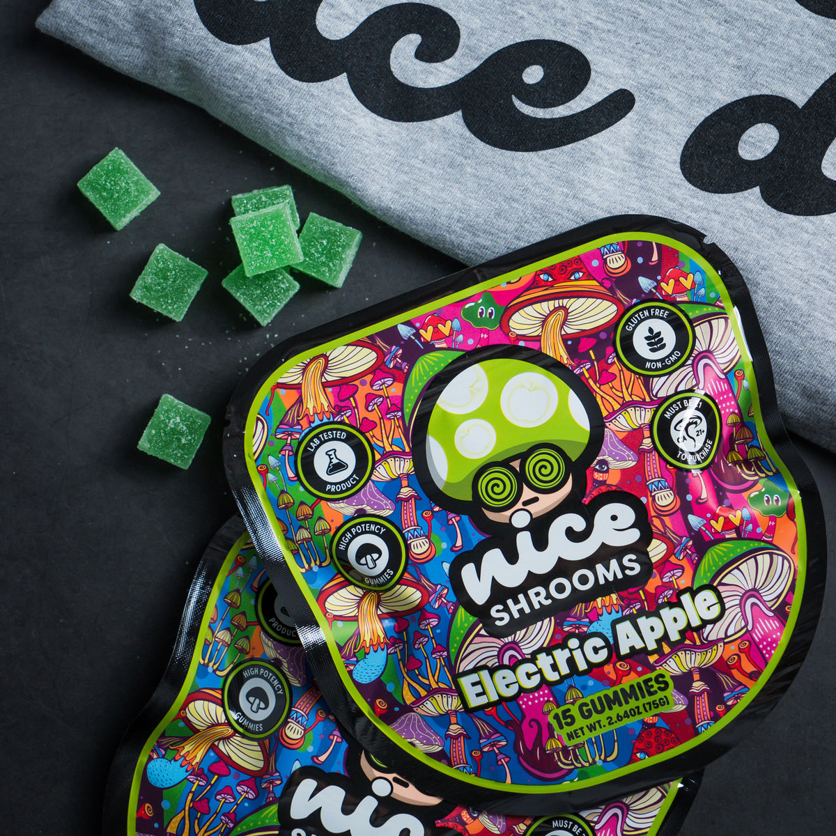 Two packages of Nice Shrooms Electric Apple gummies are shown on a dark surface next to scattered green gummies and a gray sweatshirt.
