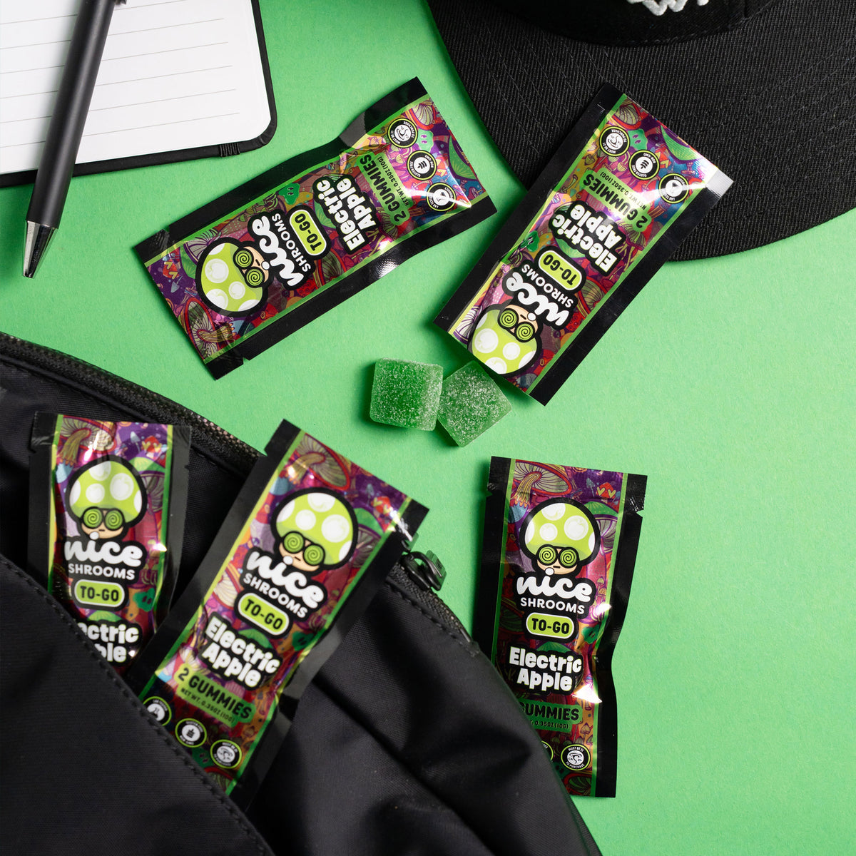 Four packets of Nice Shrooms Electric Apple bubble gummies are arranged on a bright green surface with a black zip pouch, a black cap, a pen, and a notebook nearby. Two green square gummies are placed in the center.
