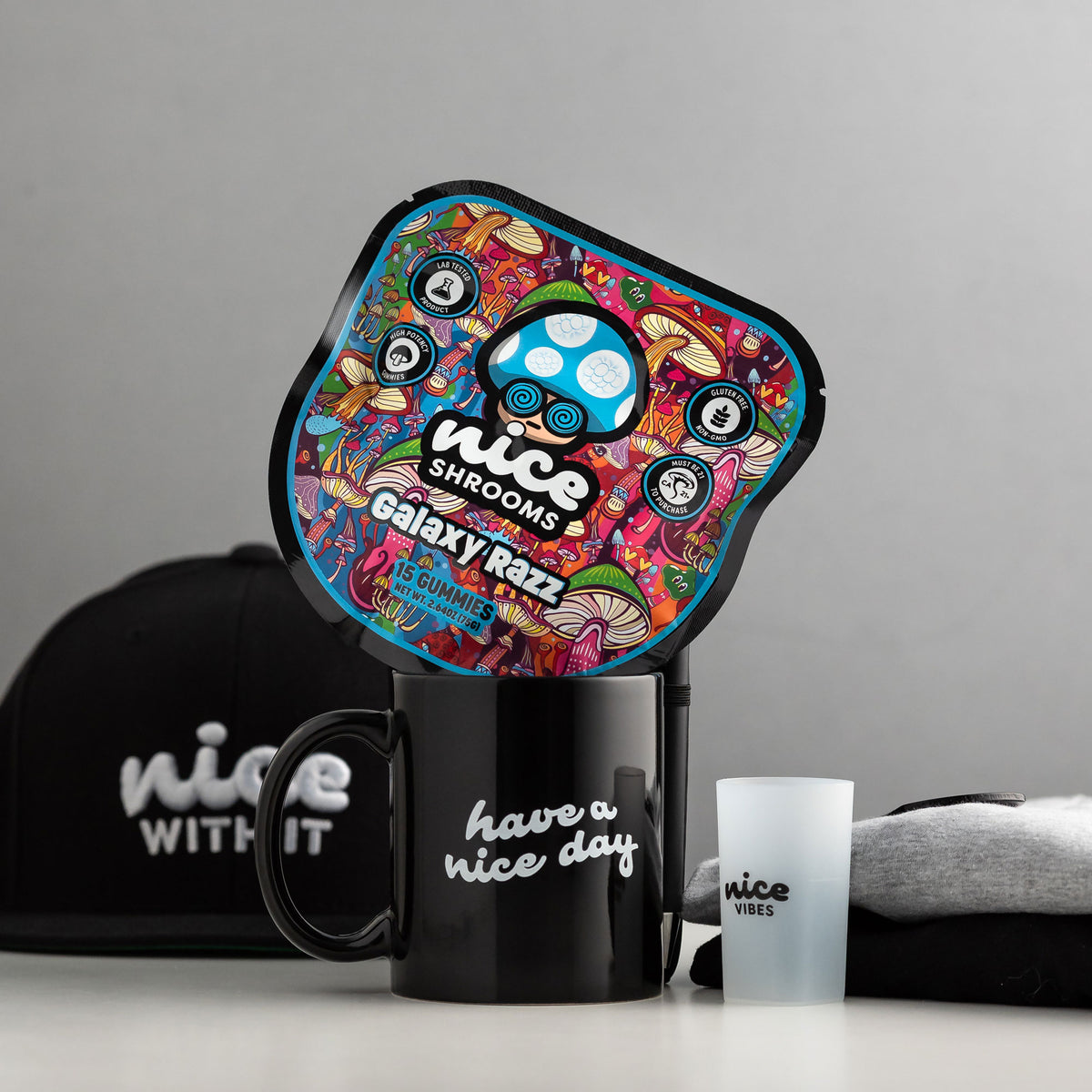 A product bundle featuring a black ceramic mug with 'have a nice day' printed in white script, a matching black baseball cap, a frosted tumbler, and a colorful mushroom-shaped coaster or mat. The main coaster features a vibrant, psychedelic design with a blue and white mushroom cap and the text 'nice SHROOMS galaxy raze'.