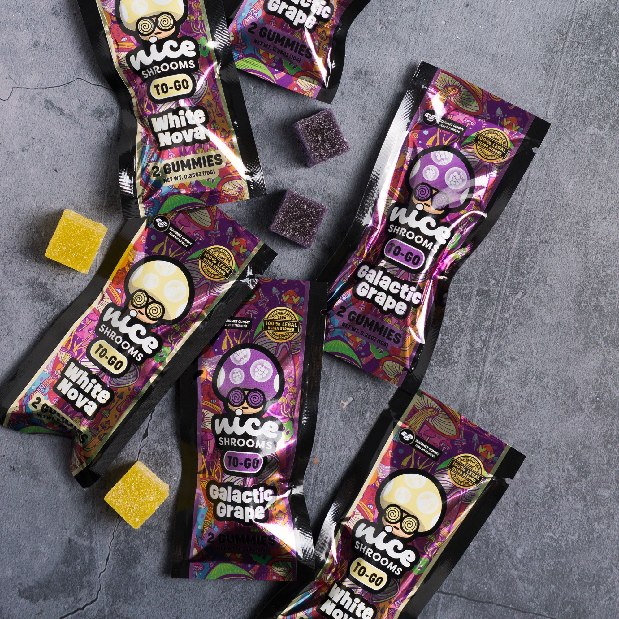 Five bags of nice shrooms gummies arranged on a gray stone-textured background, with several purple and yellow gummy cubes scattered around. The packaging features colorful psychedelic graphics in purple, pink, and black, with text for White Nova and Galactic Grape.