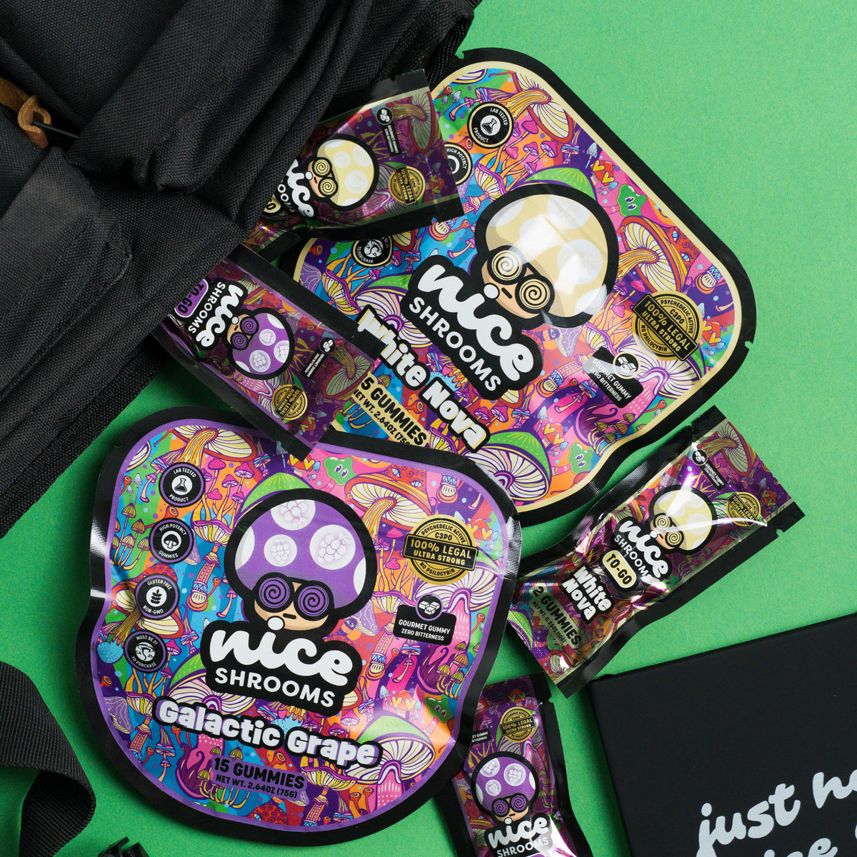 Several colorful packaged gummy candies from Nice Shrooms are arranged on a bright green surface with a black bag partially visible at the top left. The packages feature psychedelic artwork and the text 'Nice Shrooms' and 'Galactic Grape'.